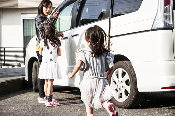 Mother and two sisters are coming to a car. Mother is beckoning to little sister. Little sister is running to her mother and elder sister standing by a car.